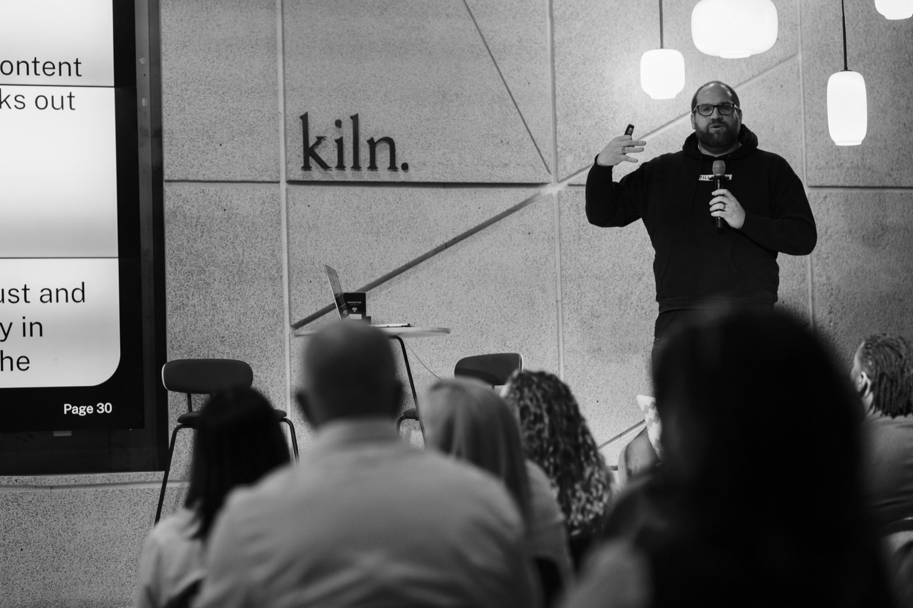 Benjamin Ard speaking on stage at kiln, holding a microphone in front of an audience.